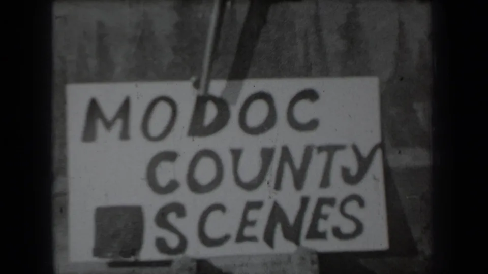 Black-and-white 1937 8mm frame showing a hand holding a revolver above a hand-lettered Modoc County Scenes title card.