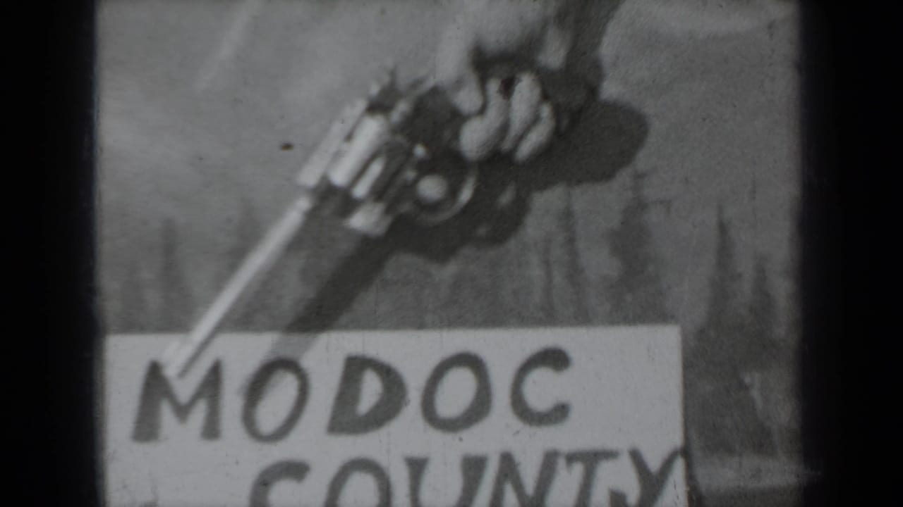 Black-and-white 1937 8mm frame with a hand gripping a revolver above the lower portion of a Modoc County title card.