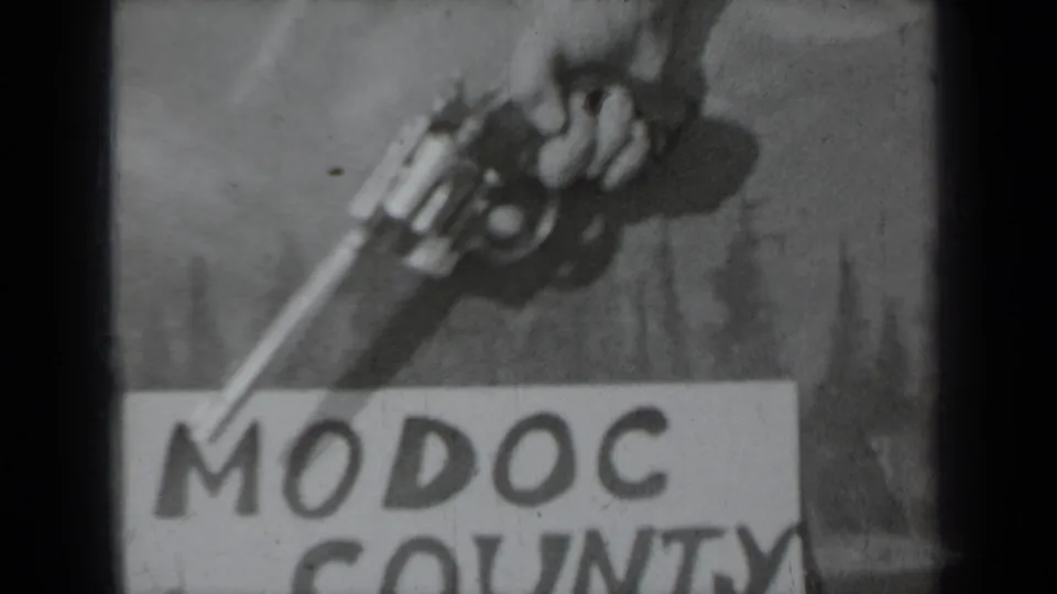 Black-and-white 1937 8mm frame with a hand gripping a revolver above the lower portion of a Modoc County title card.