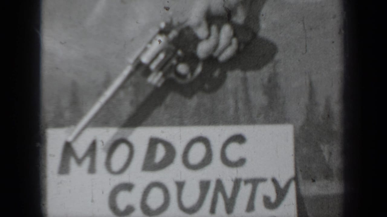 Soft black-and-white 1937 8mm frame showing a hand-held revolver angled over the words Modoc County on a paper sign.