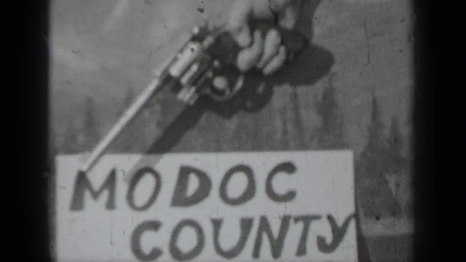 Soft black-and-white 1937 8mm frame showing a hand-held revolver angled over the words Modoc County on a paper sign.