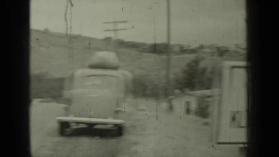 1937 8mm frame showing a car with luggage on its roof driving away along a roadside near Klamath, California, with utility poles and hills in the background.