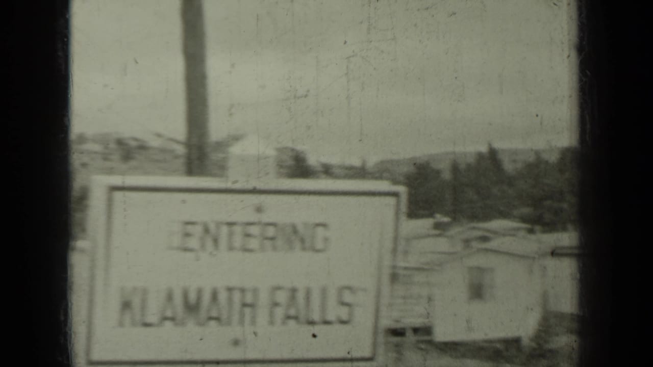 1937 8mm frame with a roadside sign reading 'ENTERING KLAMATH FALLS' near modest buildings and hills in the Klamath, California area.