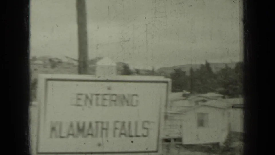 1937 8mm frame with a roadside sign reading 'ENTERING KLAMATH FALLS' near modest buildings and hills in the Klamath, California area.
