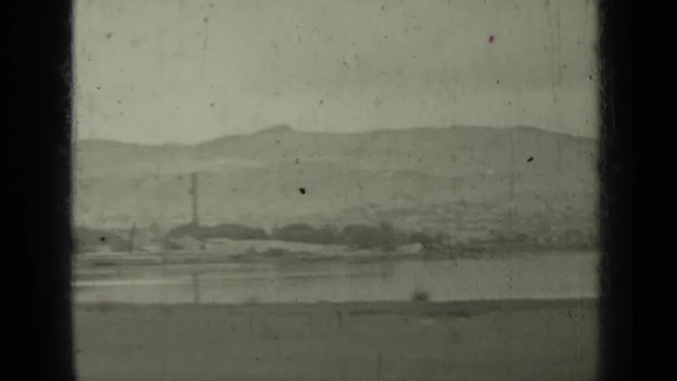 1937 Klamath-area view with a distant town, calm water, low hills, and a visible smokestack behind dark window framing.