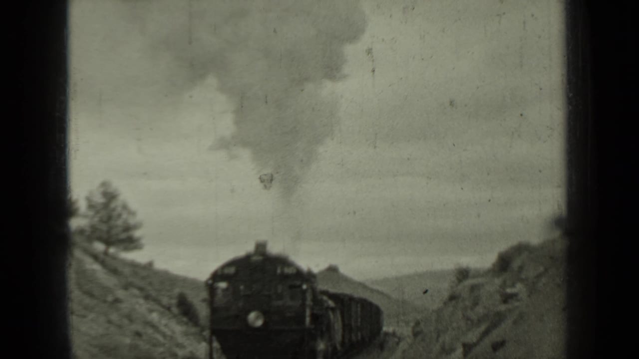 Steam locomotive approaches through a hilly rail cut with smoke above the engine.