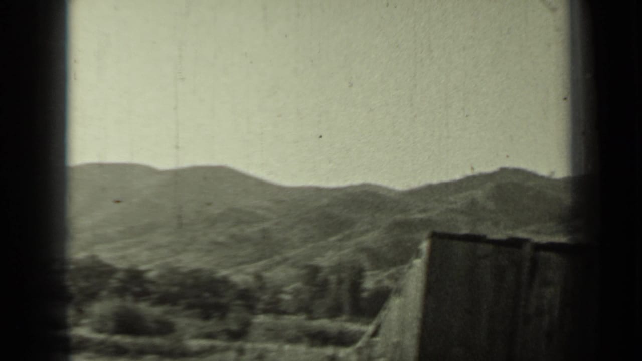 Mountain range seen through a dark window frame with fence and trees below.
