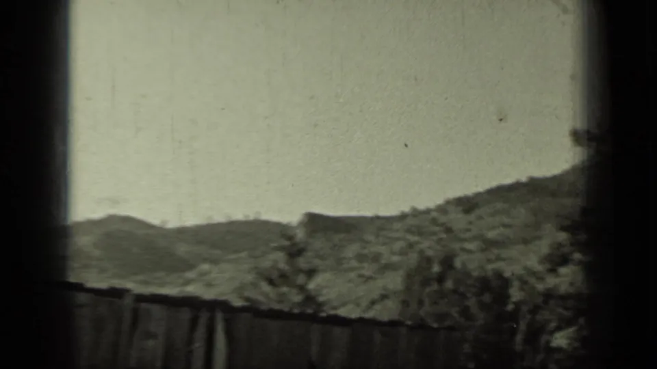 1937 Klamath, California 8mm frame with rolling hills, a rough fence, and trees partly blocked by a dark window mullion.