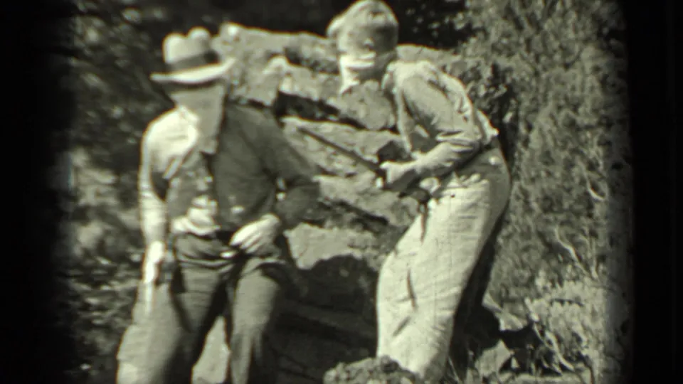 Two adult men in the 1937 Klamath clip stand by a stone wall wearing hats and cloth face coverings while holding long sticks.