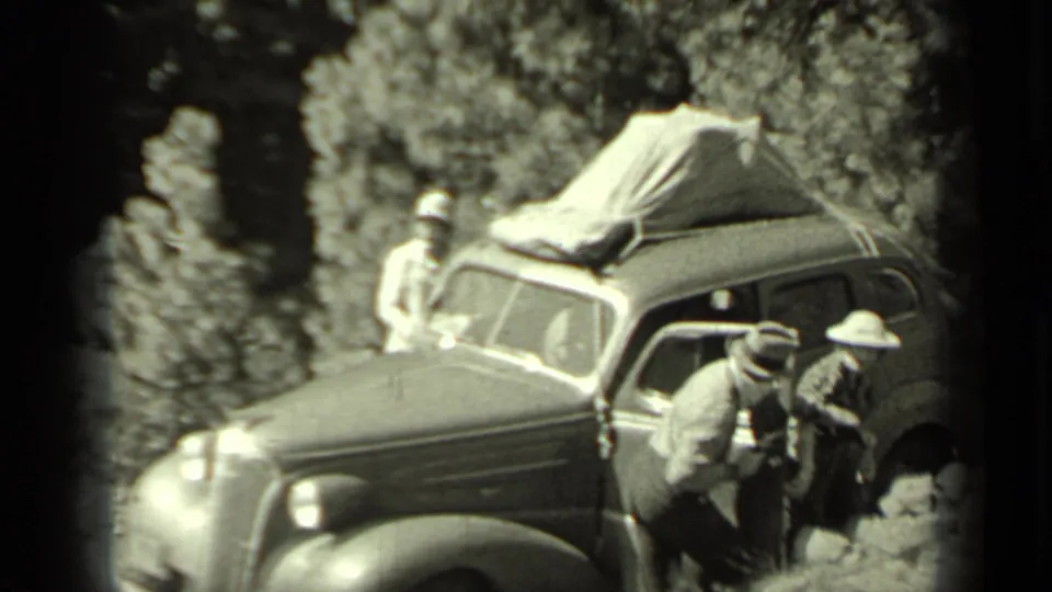 1937 Klamath, California frame showing men around a stopped sedan with canvas-covered luggage and a firearm visible in the roadside scene.