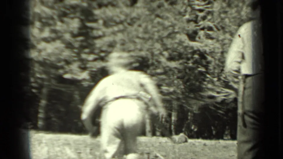 In the Klamath woods, one person bends near the ground while another figure is partly visible at the frame edge.