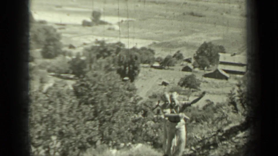Black-and-white 1937 Klamath home-movie frame showing a boy in the foreground with hillside trees, shrubs, and a distant structure behind him.