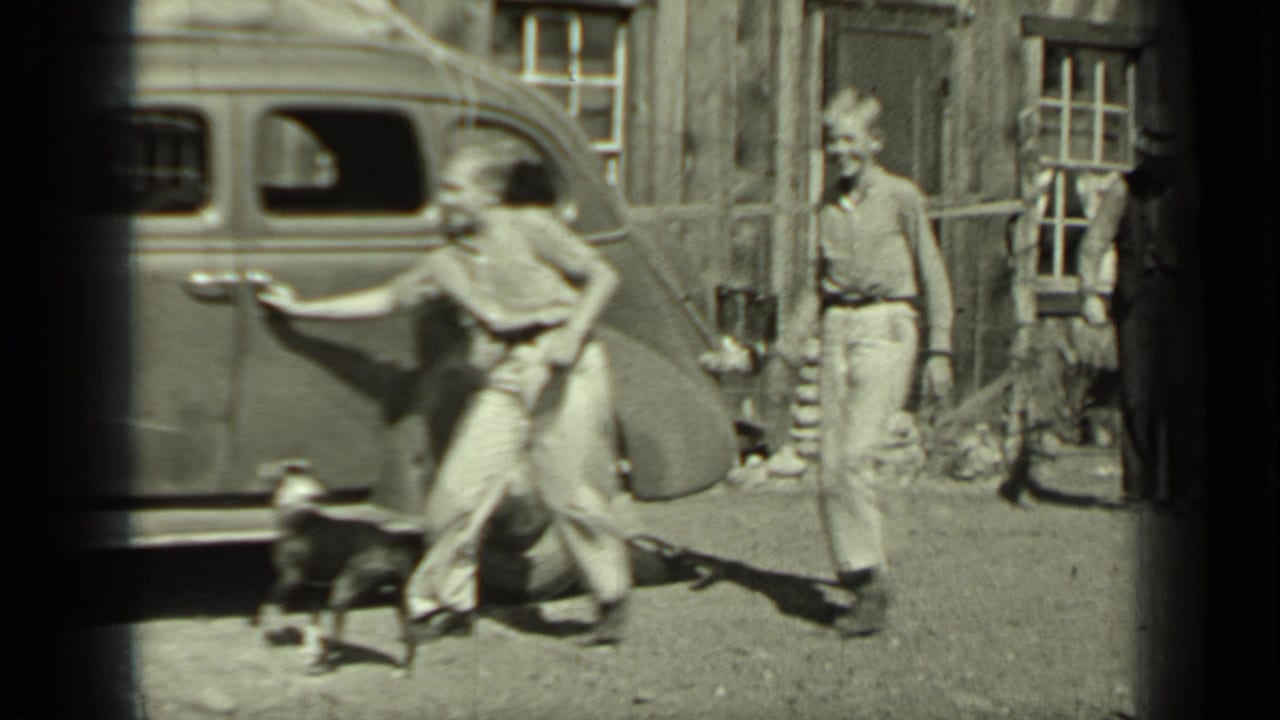 Two young people and a small dog beside a parked vintage sedan on a dirt road.