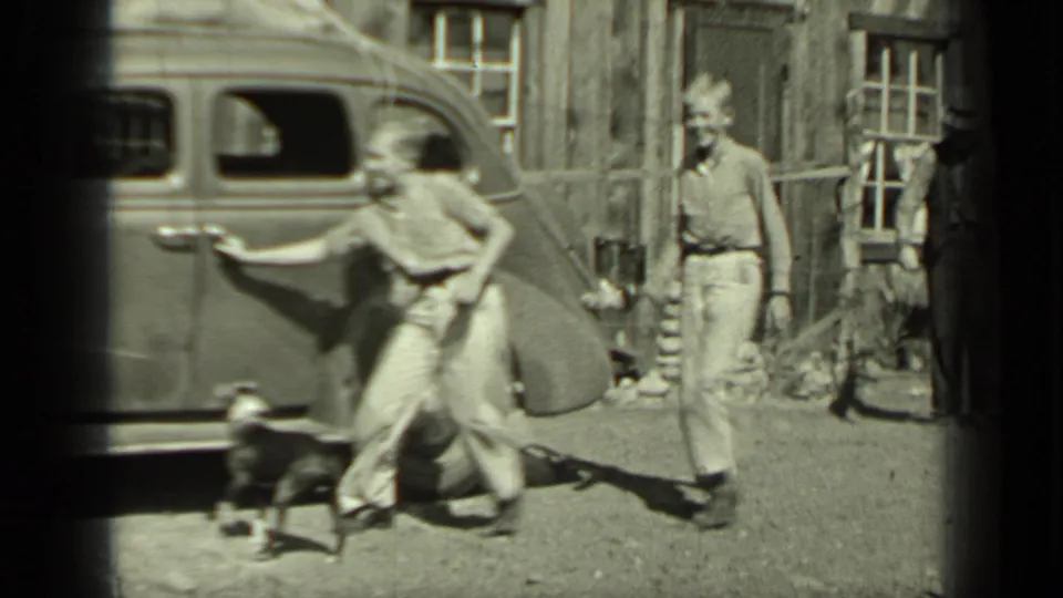 In Klamath, California, two young people move near a parked 1930s sedan while a small dog runs in front of a wood building.