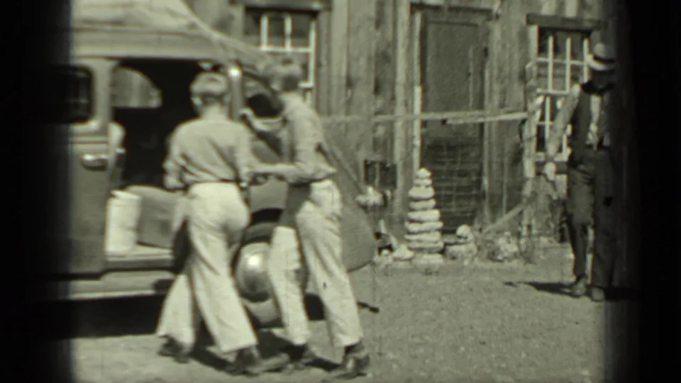 Two young people appear to hop into or lean into a 1930s sedan in Klamath, with a man standing near the wood-sided building behind them.
