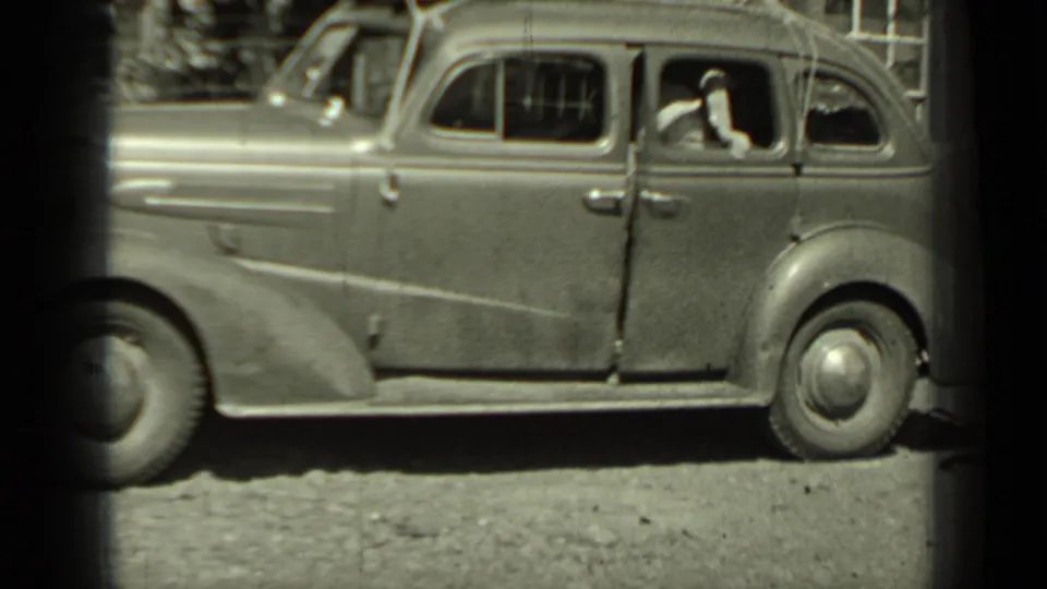 A 1930s sedan is seen from the side on a dirt road in Klamath, California, with people visible through the windows.