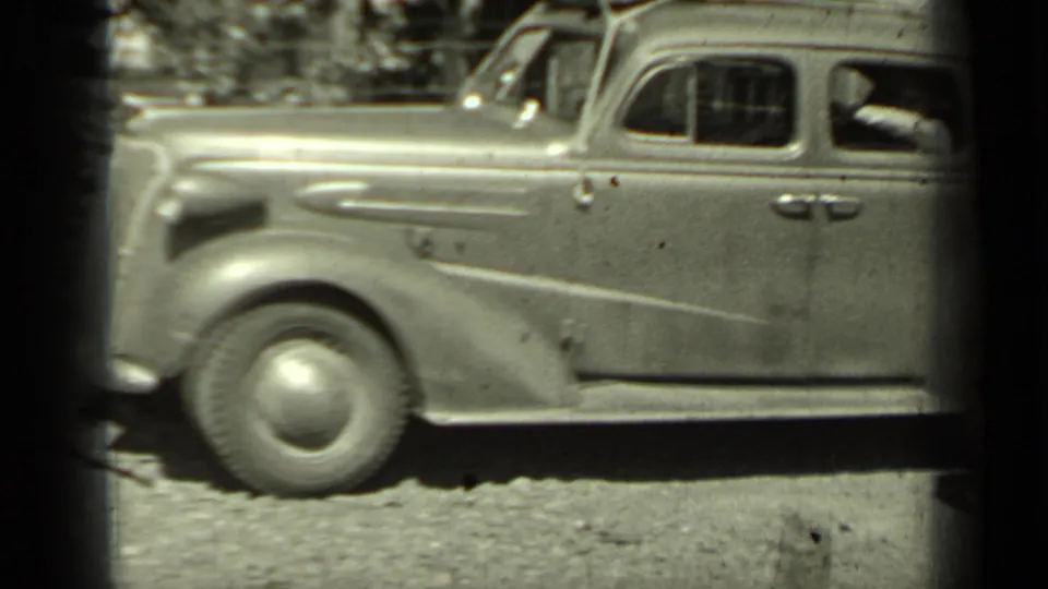 The 1930s sedan moves or is filmed in motion on the dirt road, with a passenger's arm visible through the side window.