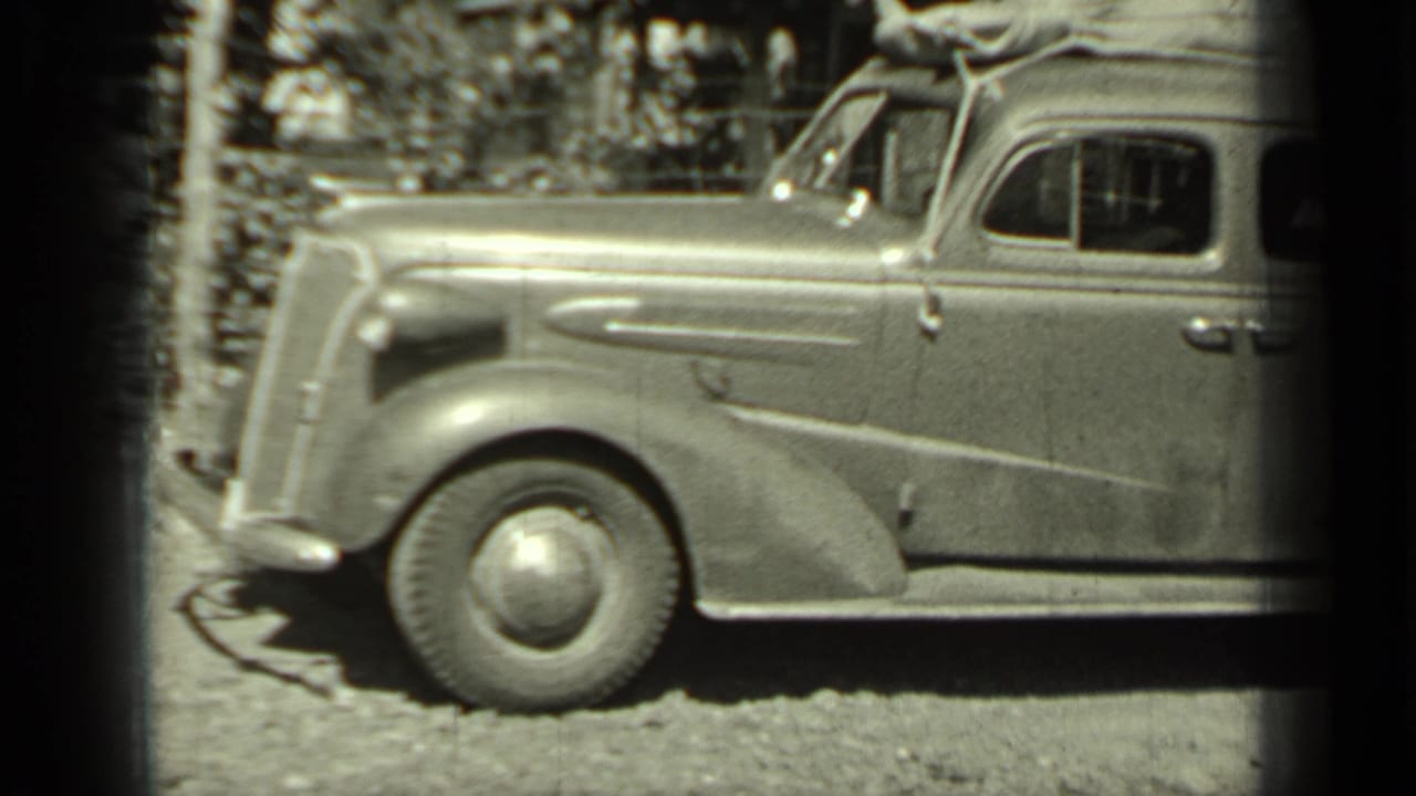 Vintage sedan parked on gravel with roof luggage and front fender visible.