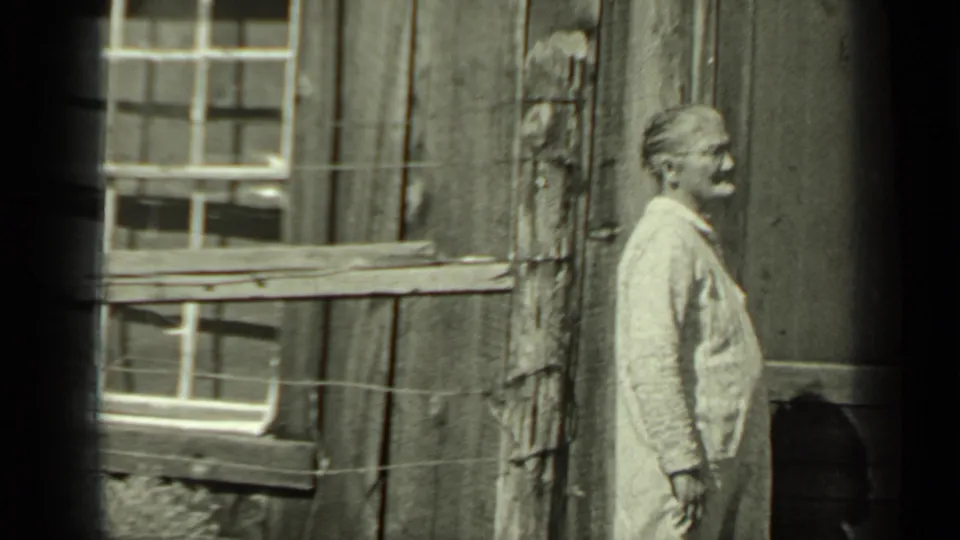 An adult stands beside a rustic wooden building and wire fence in the 1937 Klamath home-movie scene.