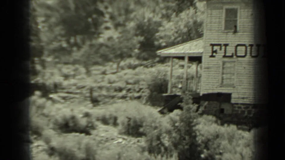 1937 Klamath, California home-movie frame showing a weathered wooden flour mill with partial FLOUR lettering beside brush and trees.