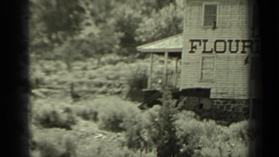 1937 8mm frame of a wooden flour mill in Klamath, California, with large painted lettering and overgrown vegetation in front.