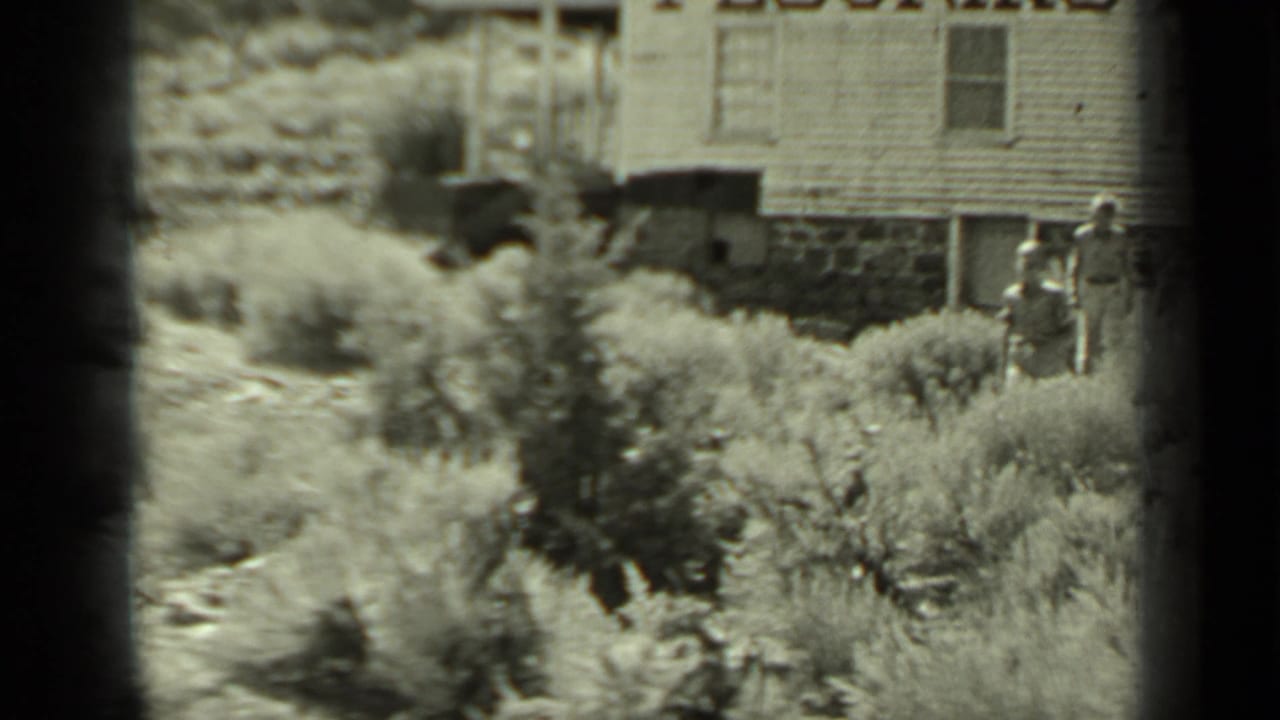 Adults walking through shrubs beside a weathered wooden building.