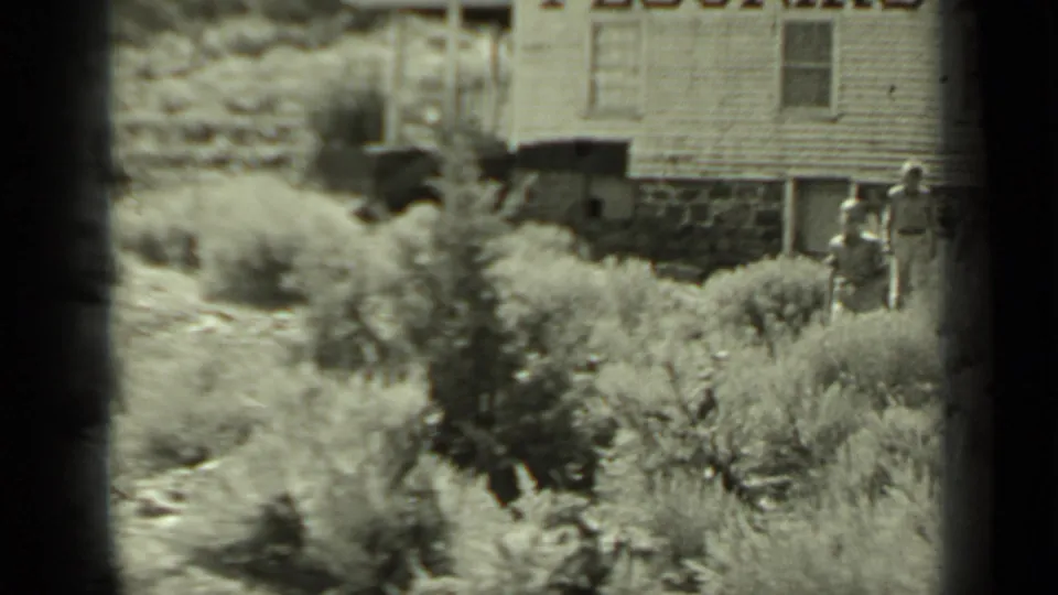 Two adults appear in dry Klamath brush near a raised wooden building, partly obscured by soft 8mm focus and frame edges.