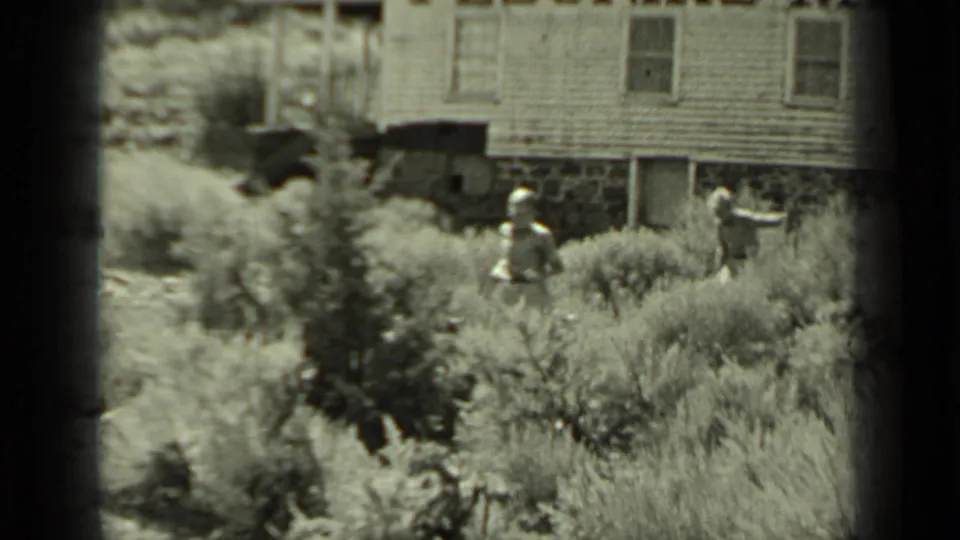 Two adults in period clothing are visible among dry shrubs below a wooden building in the 1937 Klamath home-movie scene.