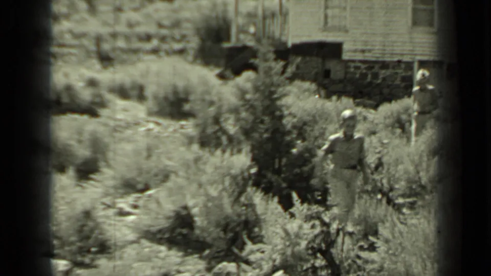 A figure stands in the brush near the wooden building as another person remains partly visible at the edge of the 1937 Klamath frame.