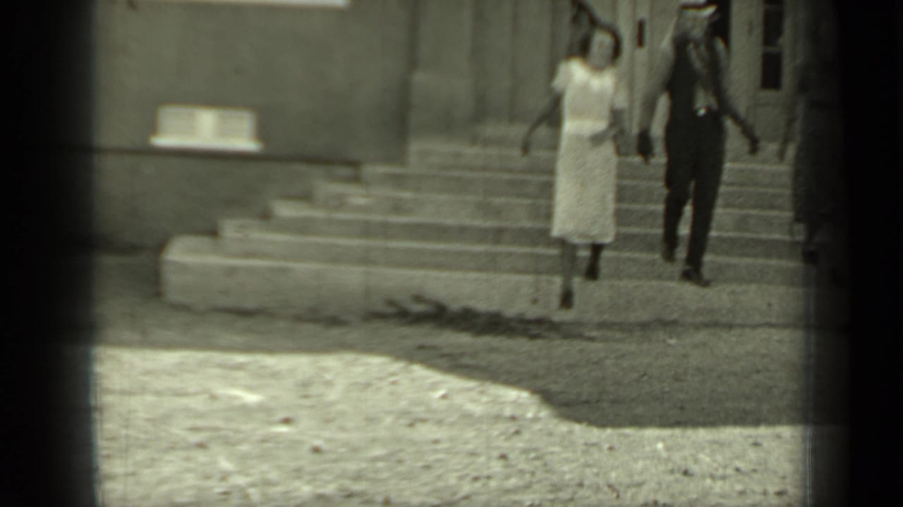 A man in a hat and vest and a woman in a light dress walk down exterior steps.