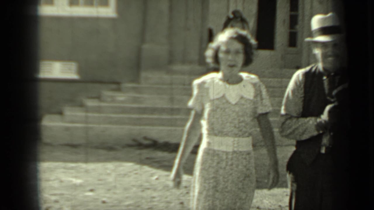 Woman in patterned dress walks near a man partly visible in a fedora by a building.