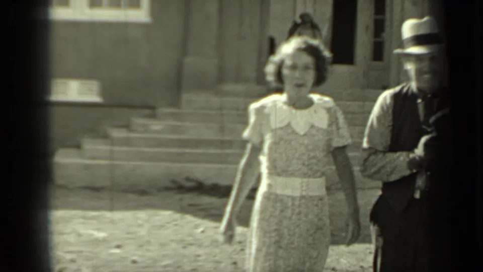 A woman in a patterned dress with a light collar walks toward the camera in Klamath, California, with a fedora-wearing man at the edge.