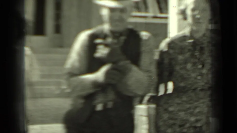 Soft 8mm frame shows the man holding a small camera and a woman beside him as they pass close to the lens outside the building.
