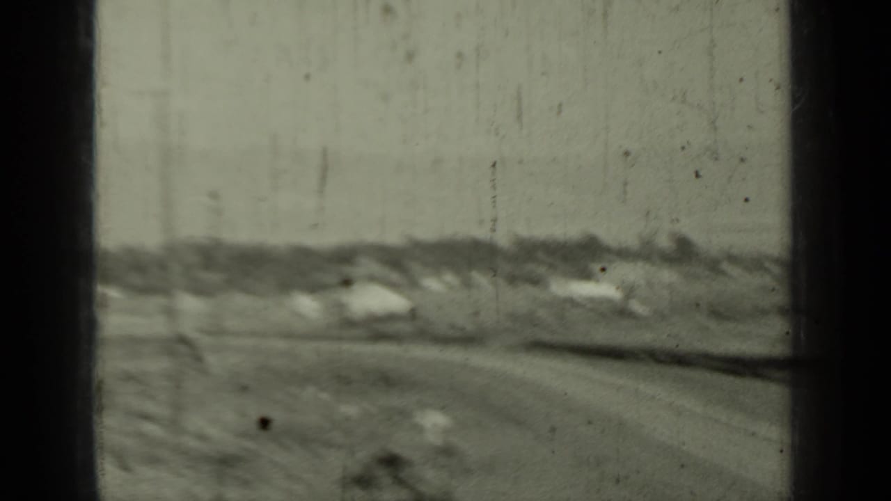 Open roadside landscape with dirt road, low buildings, brush, and soft-focus film texture.