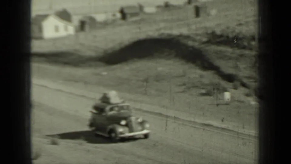 A grey 1930s car carrying roof luggage travels on a dirt road through Klamath, California, with small buildings nearby.