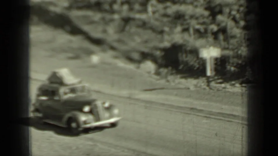 The grey car continues along a Klamath dirt road past roadside brush, utility lines, and a small sign in bright daylight.