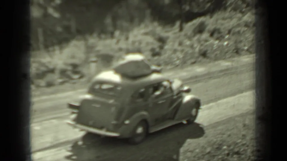 A 1937 Klamath road-trip frame shows a vintage car with roof luggage moving along a wooded road in black-and-white 8mm footage.
