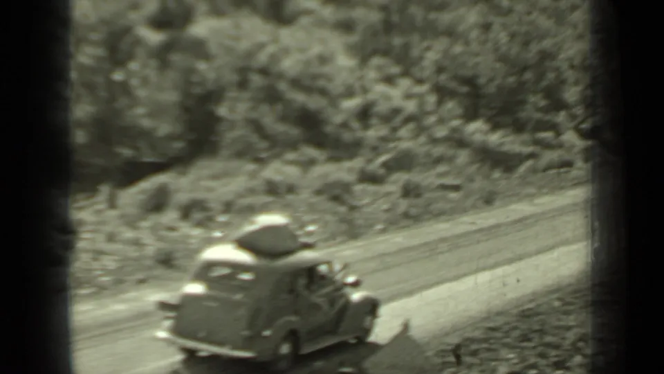 The car with roof luggage recedes along a tree-lined Klamath road, framed by soft grain, sunlight, and dark 8mm gate edges.