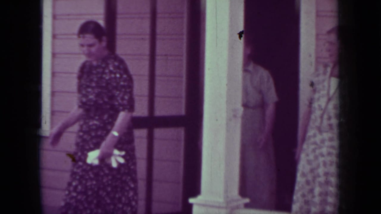 Woman in a dark floral dress walks beside a porch and white column.