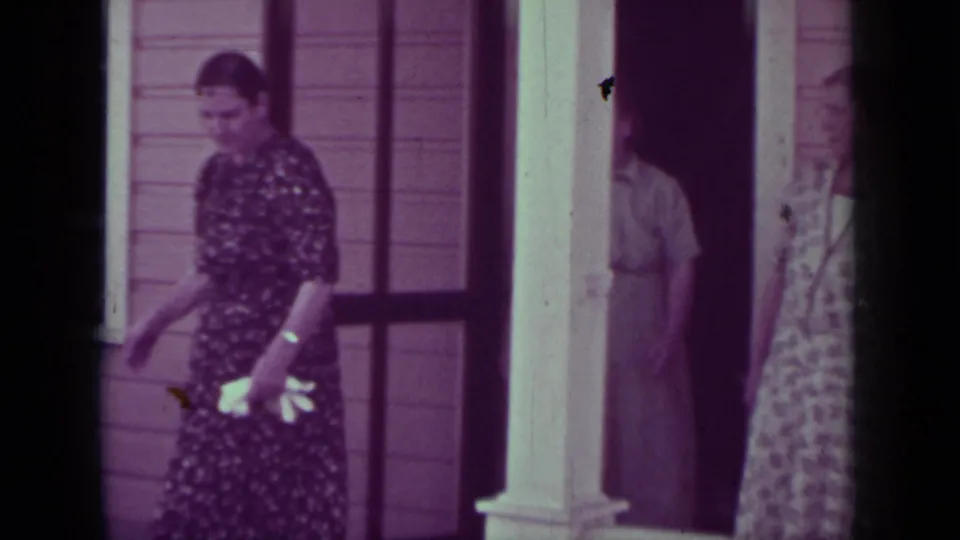 In 1937 Medford, Oregon home-movie footage, a woman in a dark floral dress walks away from a porch with wooden siding behind her.