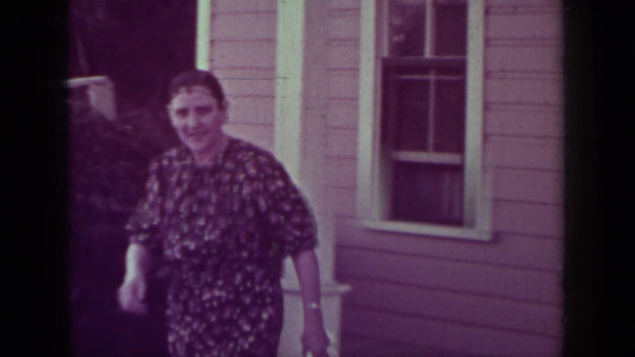 Woman in a floral dress walks near a house while others stand in the doorway.