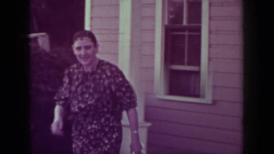 A woman in a dark floral dress walks toward the camera outside a Medford house, with two other women partly visible in the doorway.
