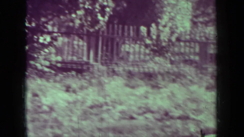 1937 8mm frame from Medford, Oregon showing a fenced yard with overgrown garden plants and a barely visible adult at the left edge.