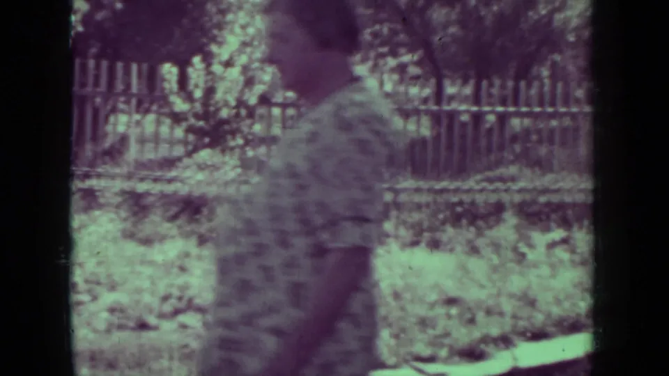 A softly focused adult figure walks in profile past a fenced garden area in the 1937 Medford, Oregon home-movie scene.
