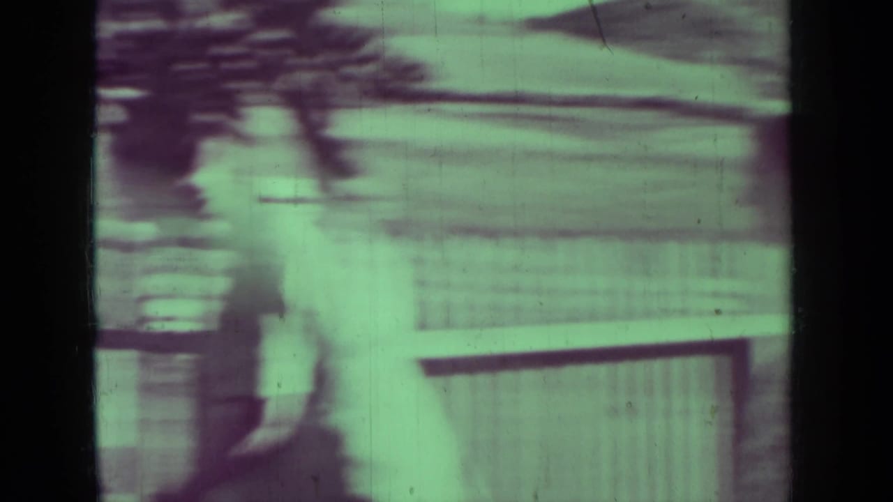 Green-tinted 8mm home-movie frame from the Medford, Oregon clip showing a blurred fence or structure beside a quiet roadside.