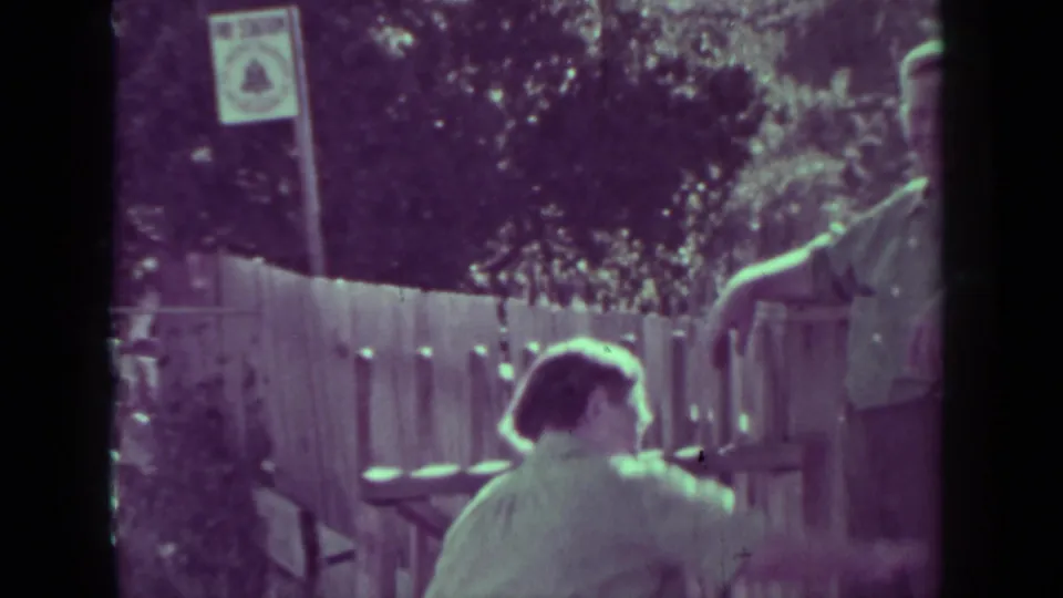1937 Medford, Oregon home-movie frame showing two men beside a wooden fence with trees and a posted notice nearby.