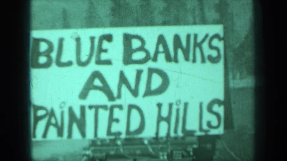 Close 1937 Medford, Oregon frame of a hand-painted Blue Banks and Painted Hills sign, softly focused in green-tinted 8mm footage.