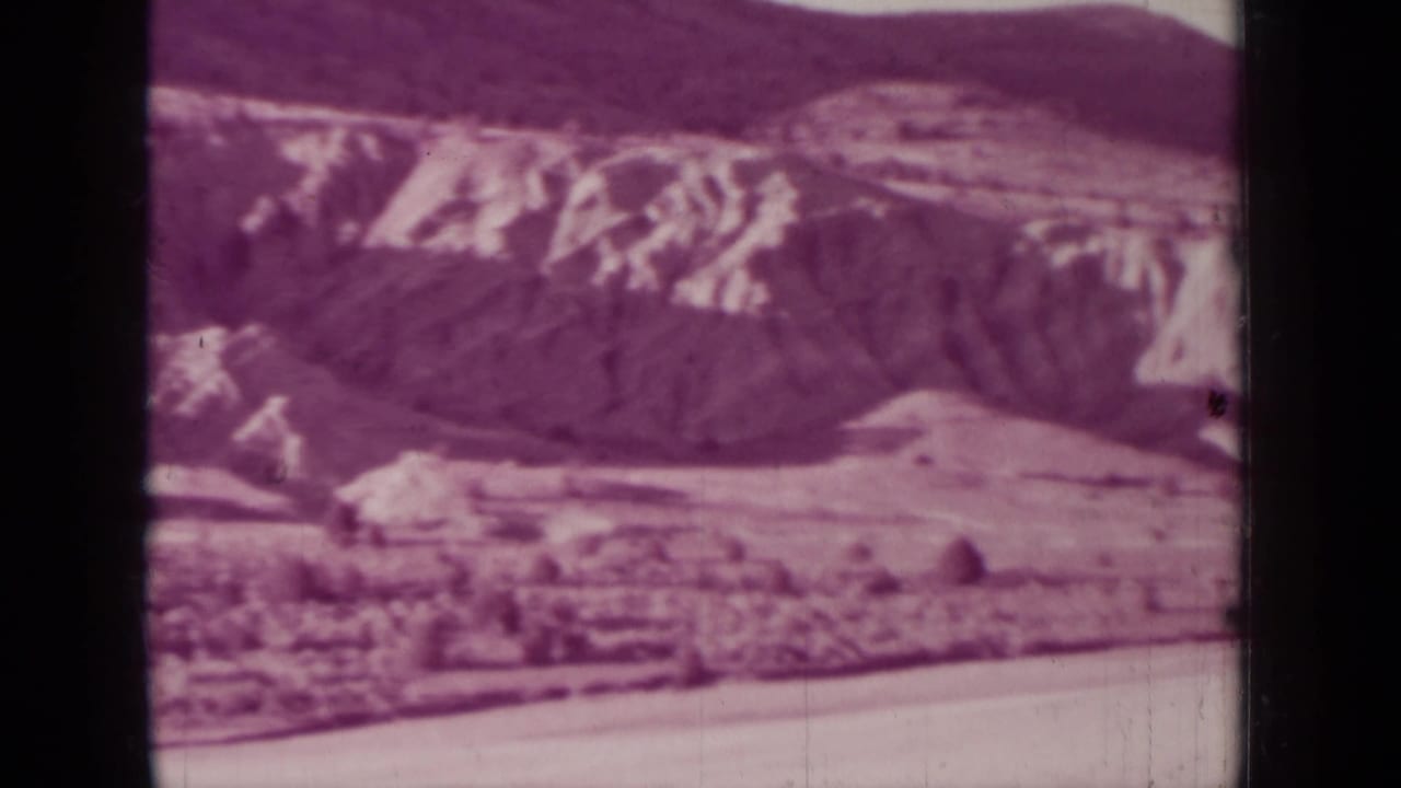 Soft 8mm frame of layered hills, a valley floor, and dark ridges under a pale sky.