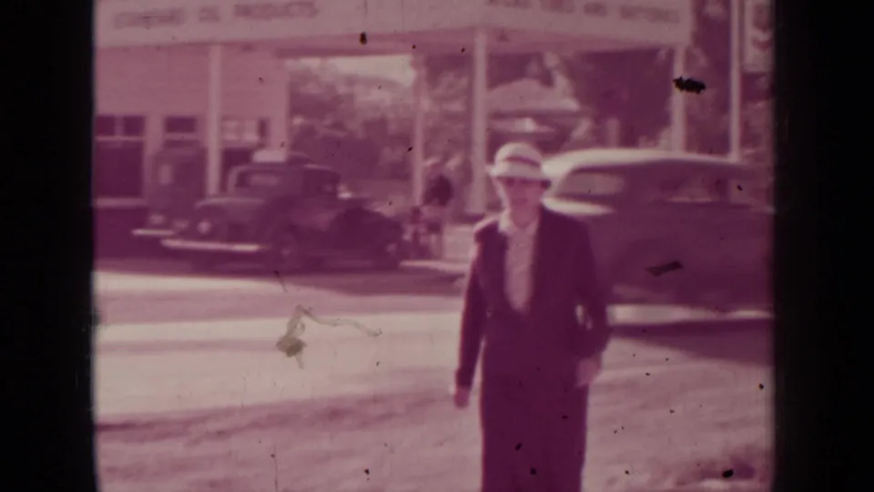 The woman continues toward the camera on a Medford street, with 1930s automobiles and commercial signage in the background.