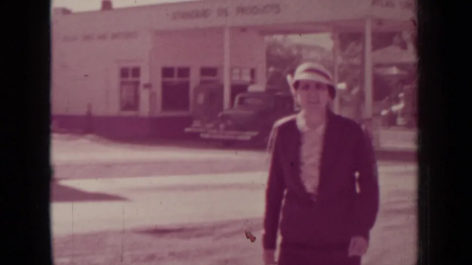 A closer frame shows the woman walking toward the camera, wearing a brimmed hat, dark suit jacket, and light blouse.
